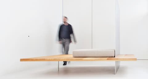 Man stepping across a low wooden platform with cushion by Roger Stüssi in a white room