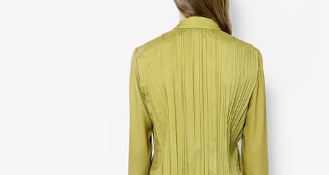 Back view of a woman in a yellow-green pleated jacket against a white background