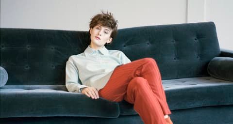 Model in a pale shirt and rust-red trousers reclining on a dark green sofa, Studio Winkler