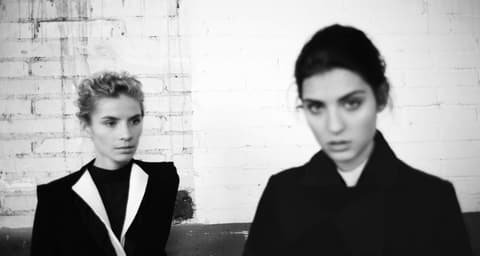 Black and white photo of two models in dark Claudia Zuber outfits in front of a white brick wall