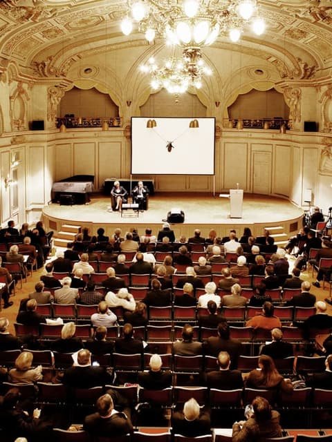 Full auditorium at Fondsmesse Schweiz: audience facing a stage with a screen beneath a chandelier