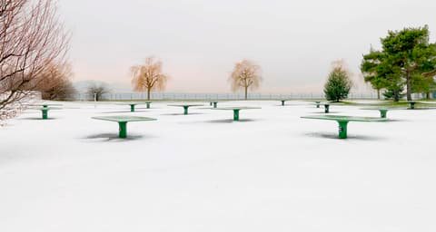 Snow-covered park with green tables and bare trees, photographed by Luxwerk