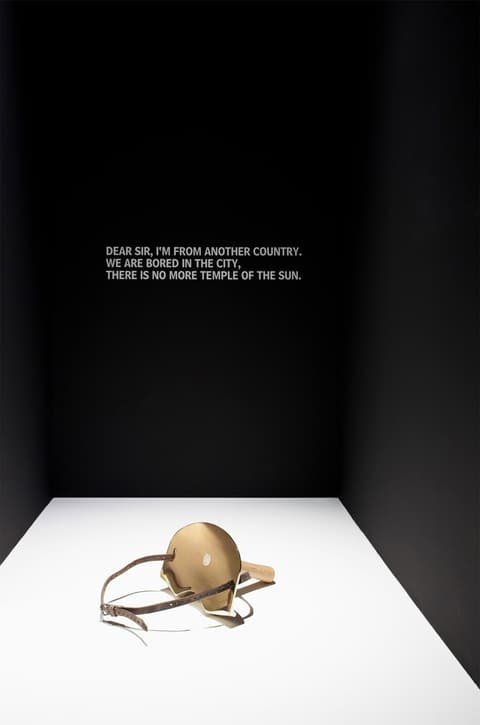 Brass and wood milking stool with leather strap by Roger Stüssi in a black exhibition niche with wall text